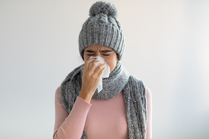 woman in sweater, hat, and scarf blowing nose into tissue