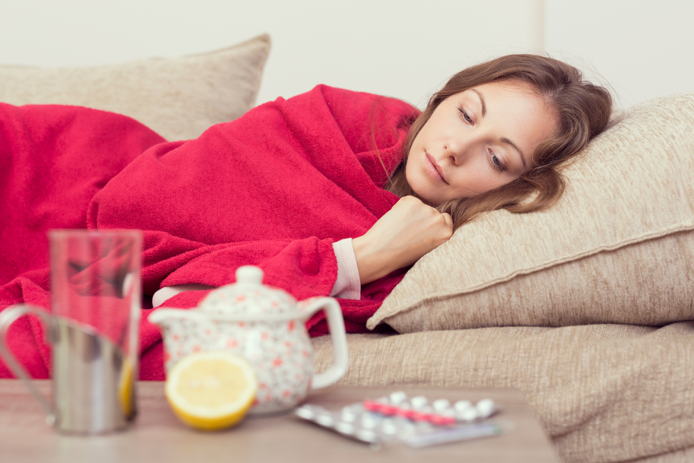 sick woman covered with a blanket lying on couch next to water, teapot, lemon, and medication pouches